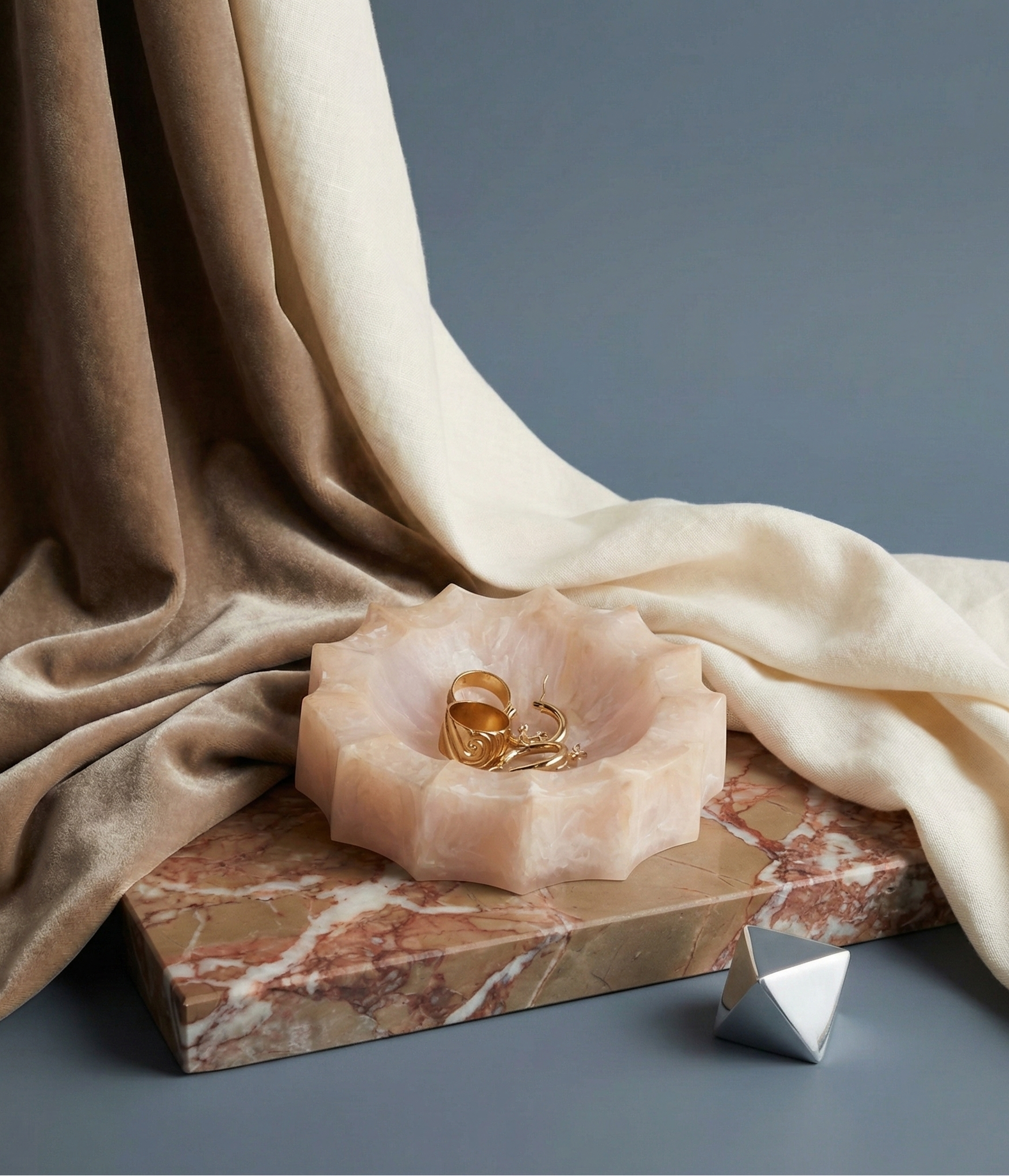 Rose quartz flower-shaped dish with gold rings on a marble base, draped with fabric against a blue background.