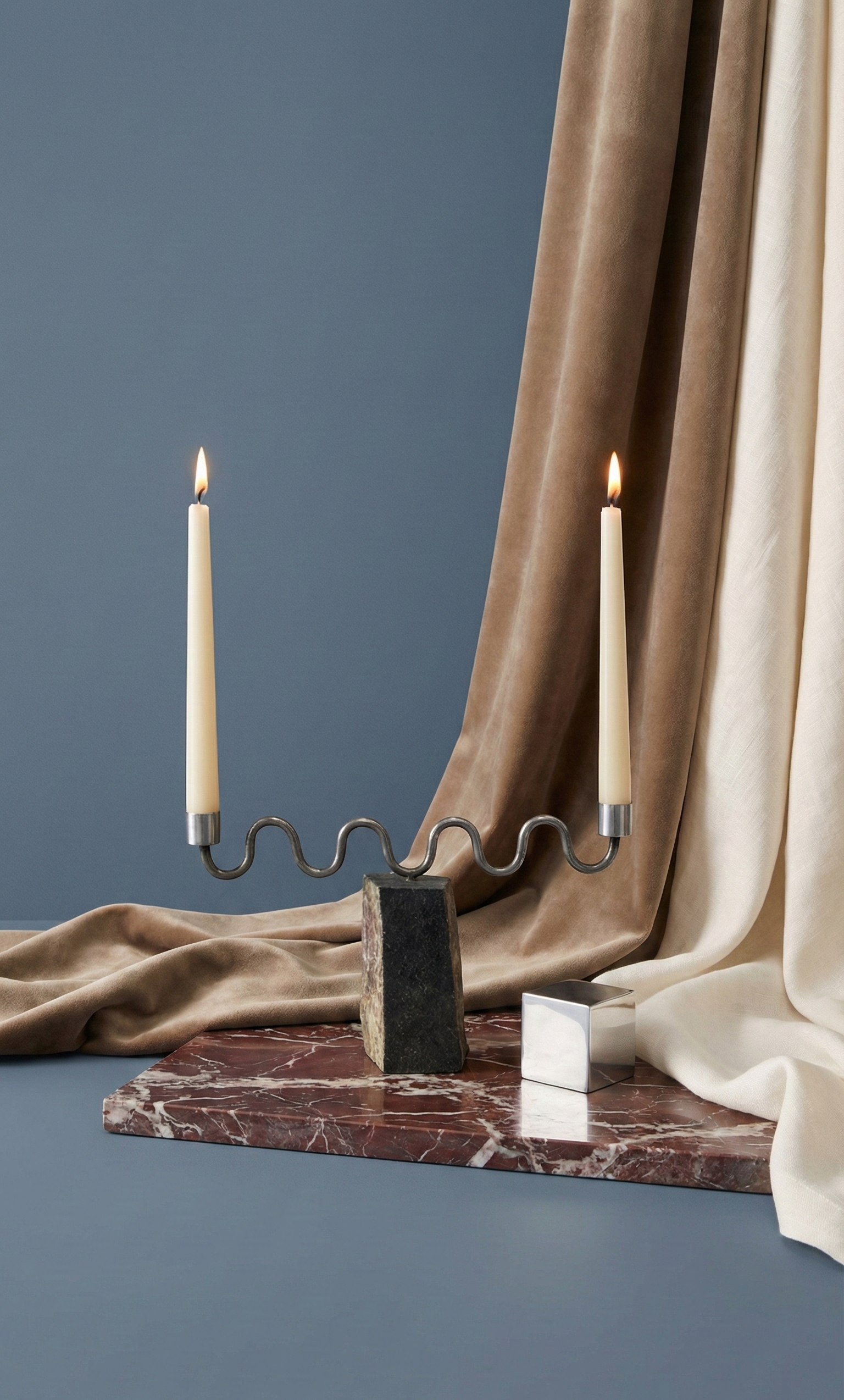 Candle holder with two candles on a marble slab against a blue wall with draped fabric.