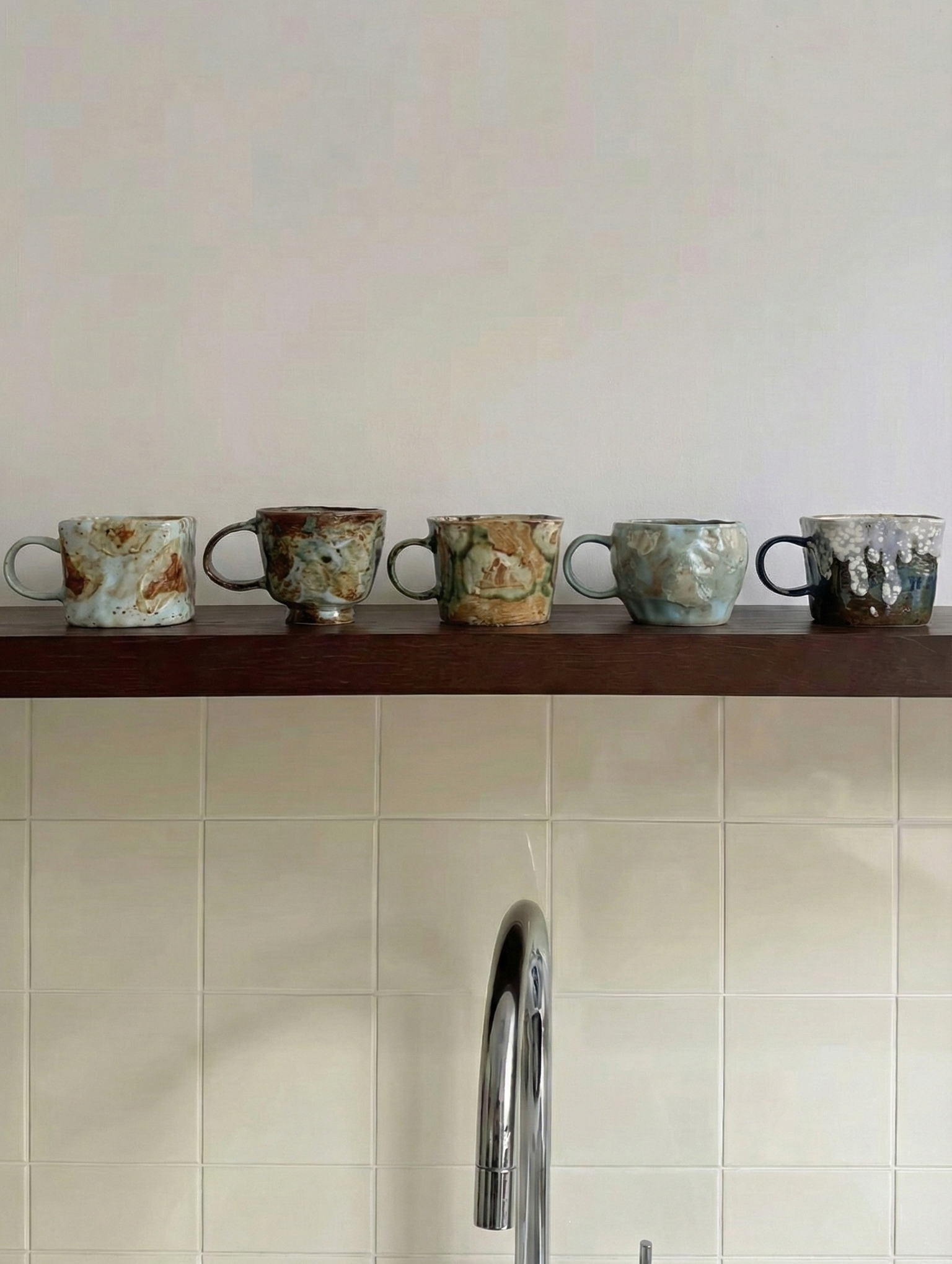 Set of ceramic cups on a wooden shelf against a tiled wall with a kitchen faucet.