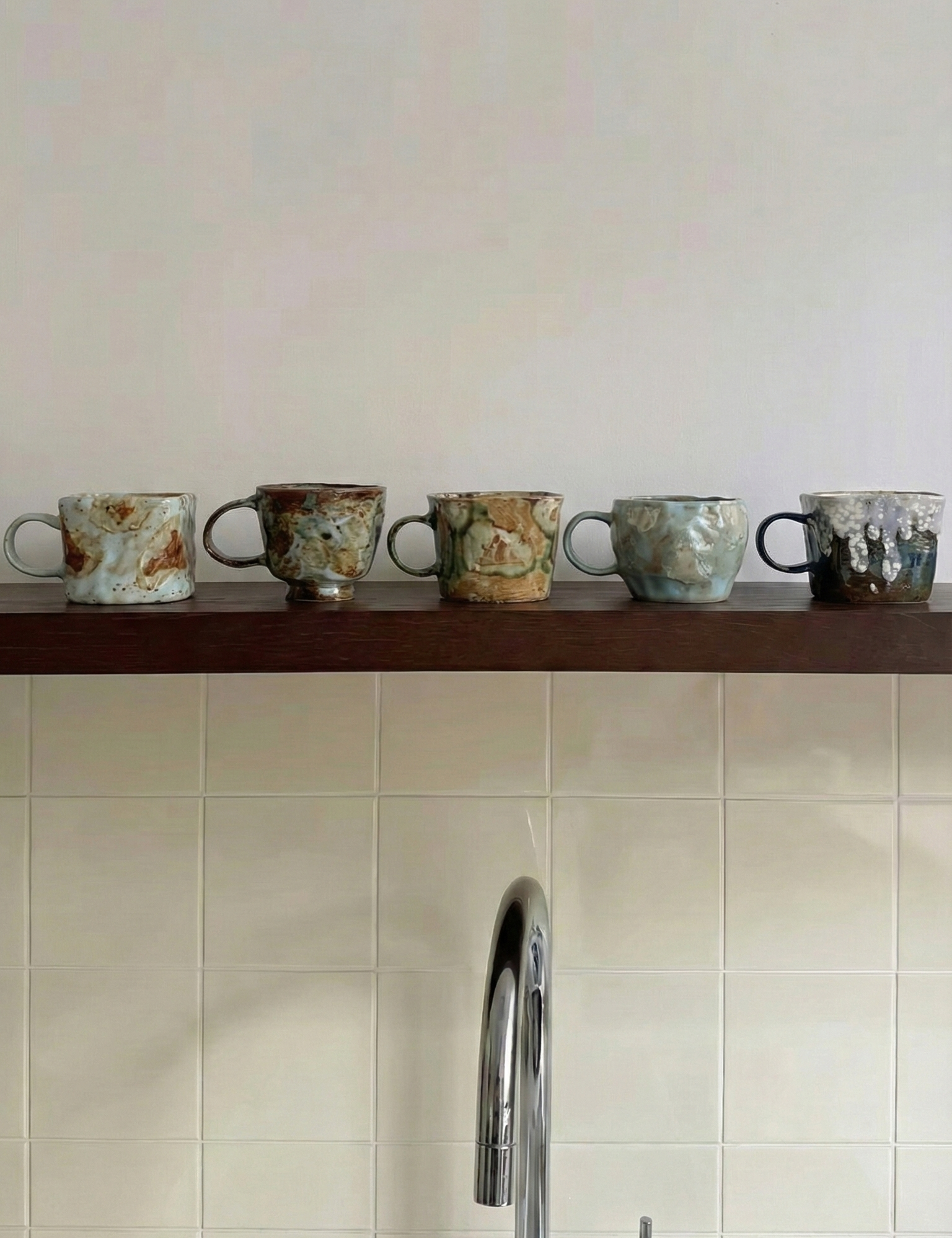 Set of ceramic cups on a wooden shelf against a tiled wall with a kitchen faucet.