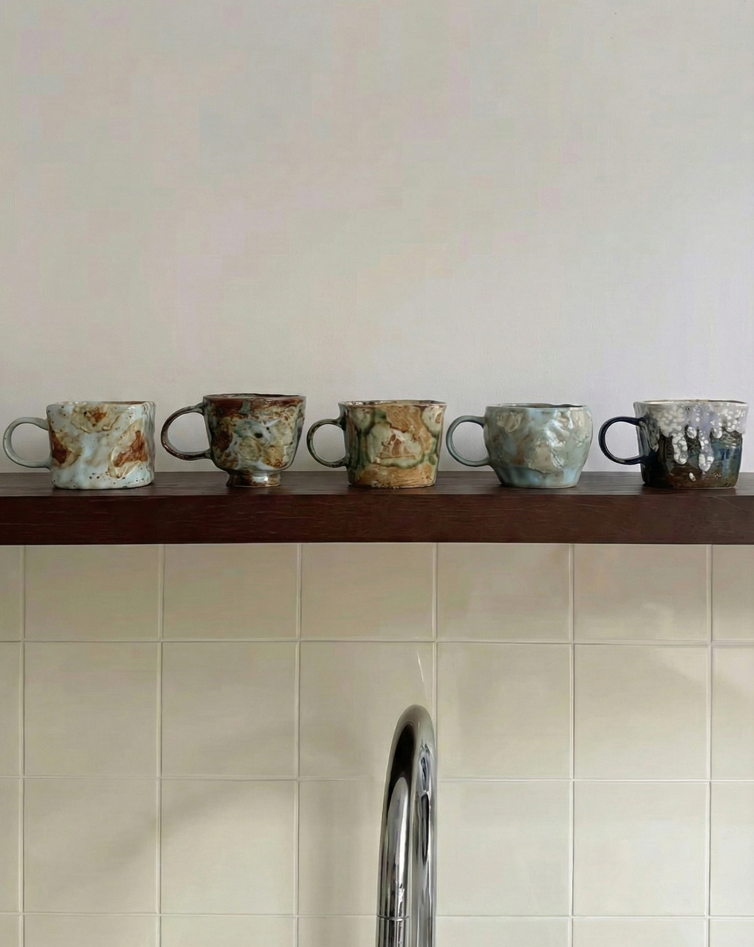 Set of ceramic cups on a wooden shelf against a tiled wall with a kitchen faucet.