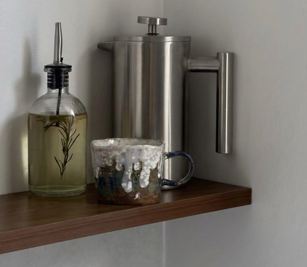 Wooden shelf with a glass bottle, metal container, and ceramic mug against a light gray wall.