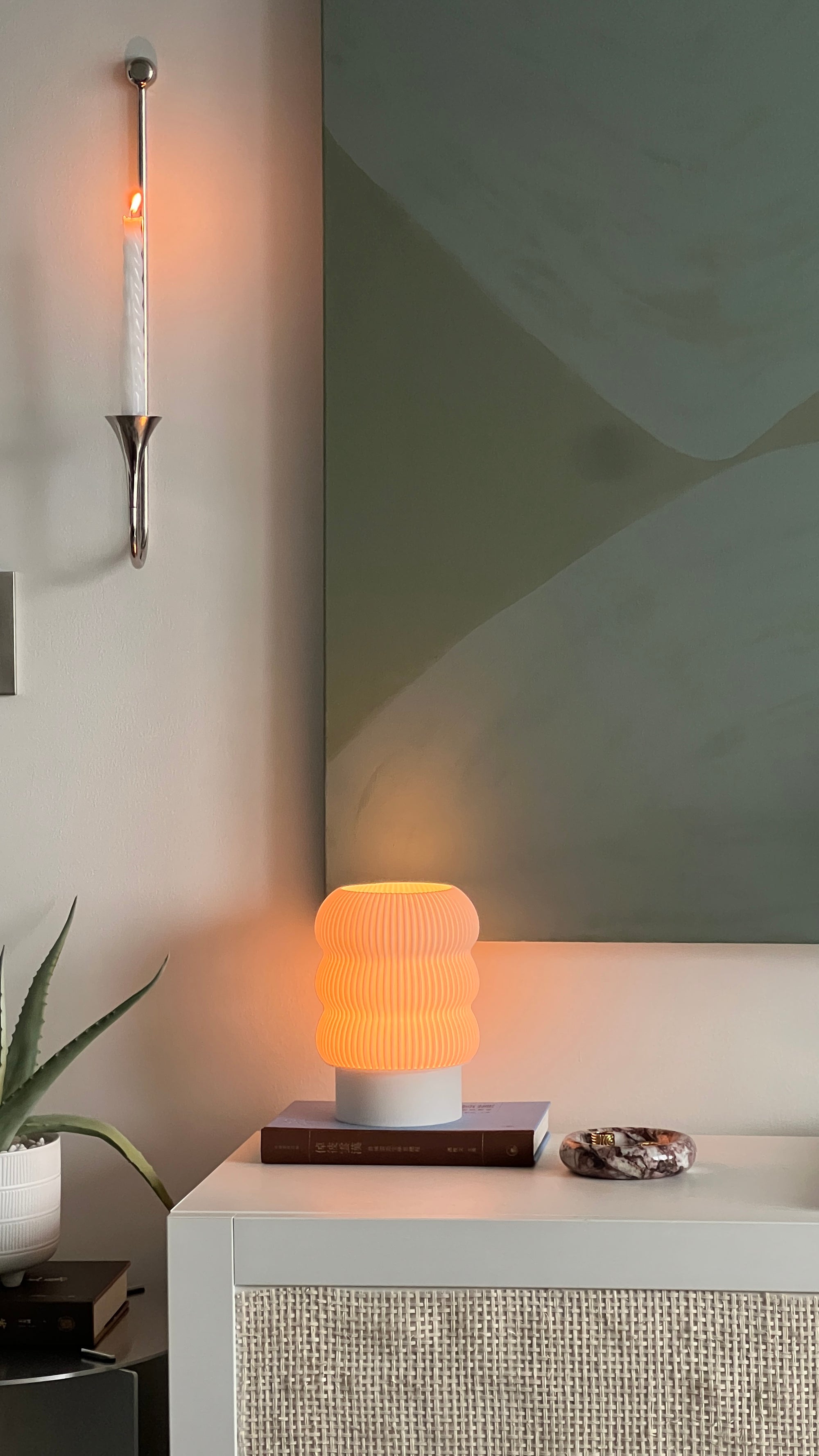 Table lamp with warm glow on a white surface next to a plant, with a wall-mounted light fixture in the background.