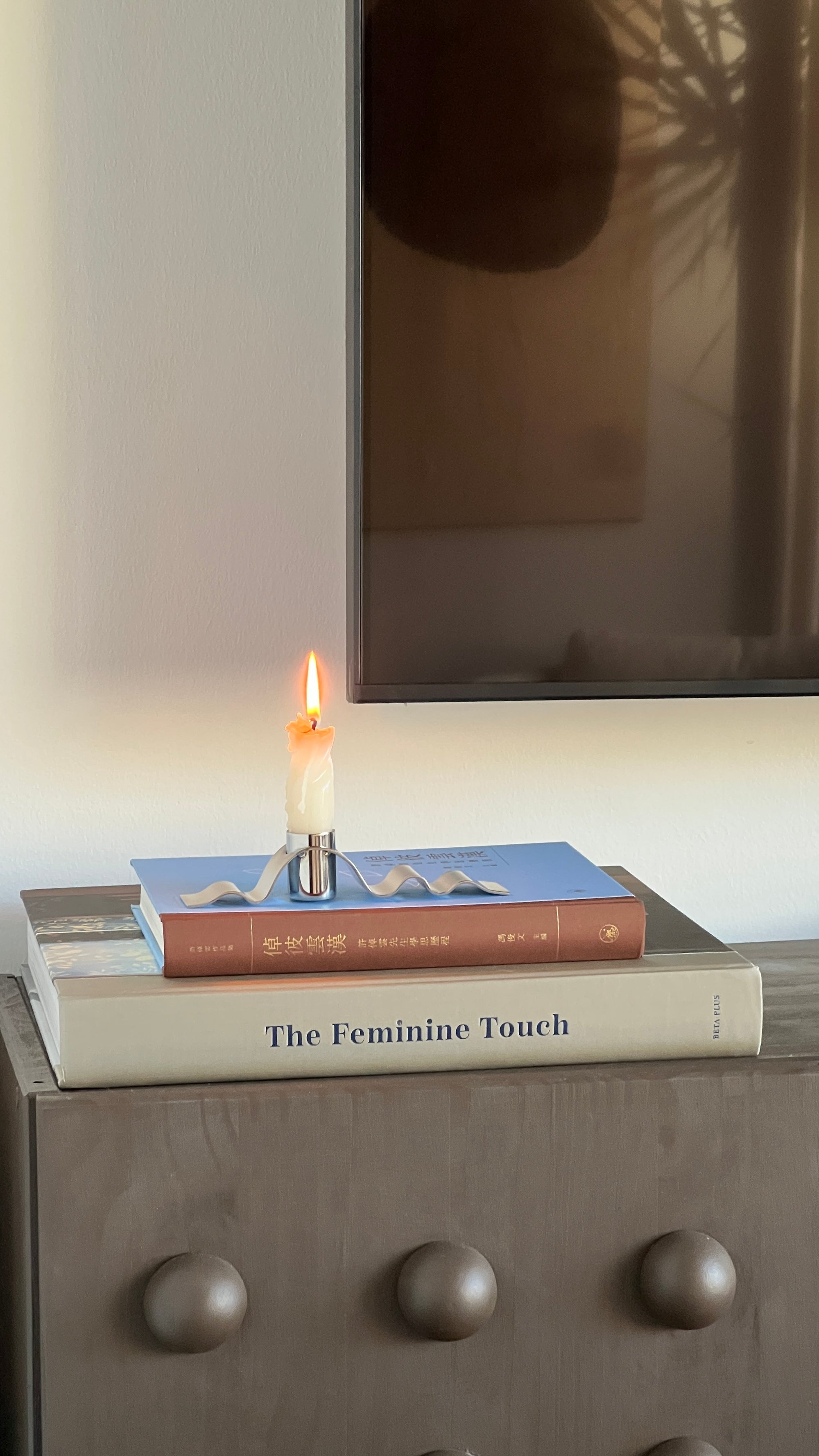Candle on books titled 'The Feminine Touch' with a reflective surface in the background