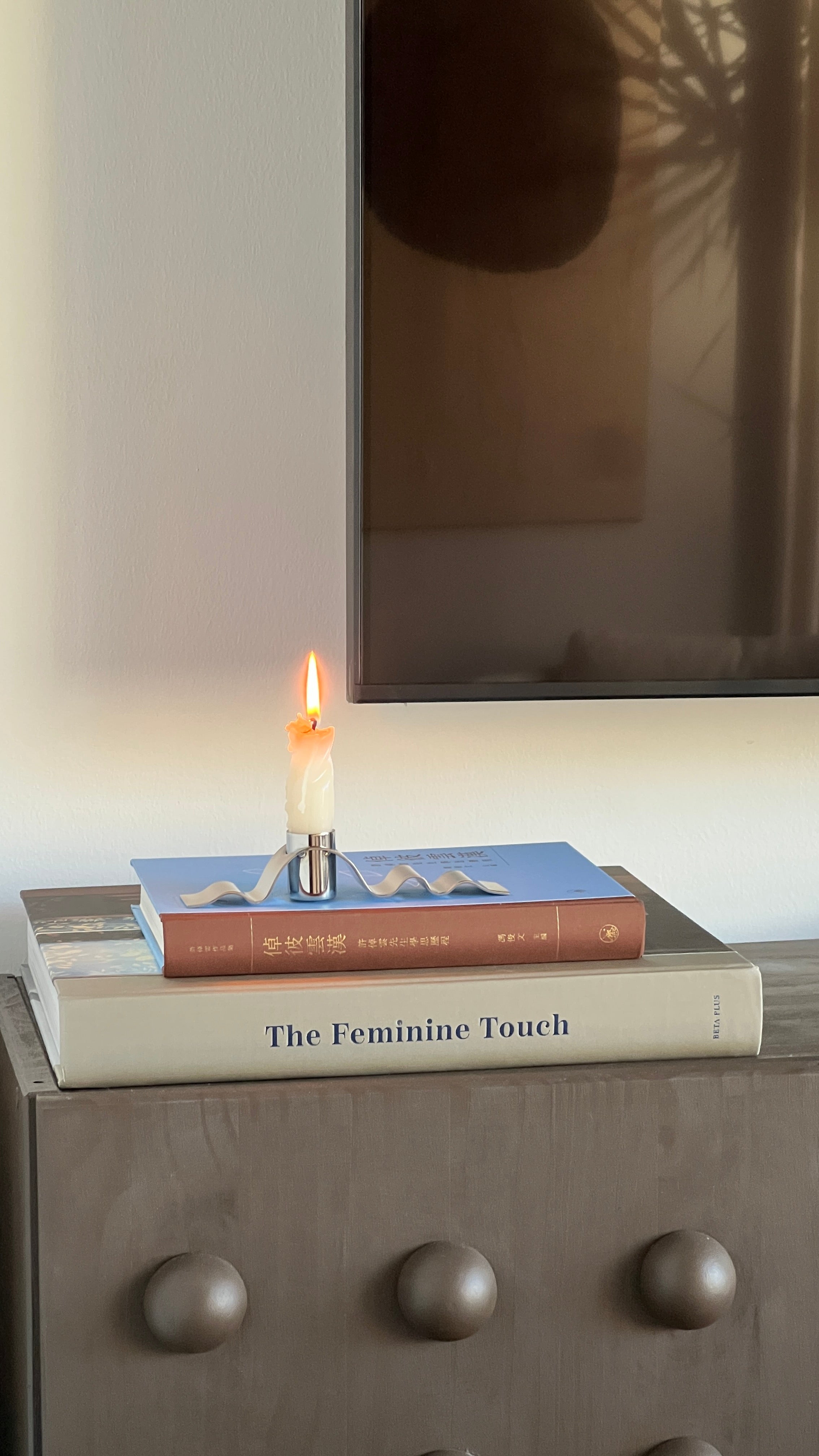 Candle on books titled 'The Feminine Touch' with a reflective surface in the background