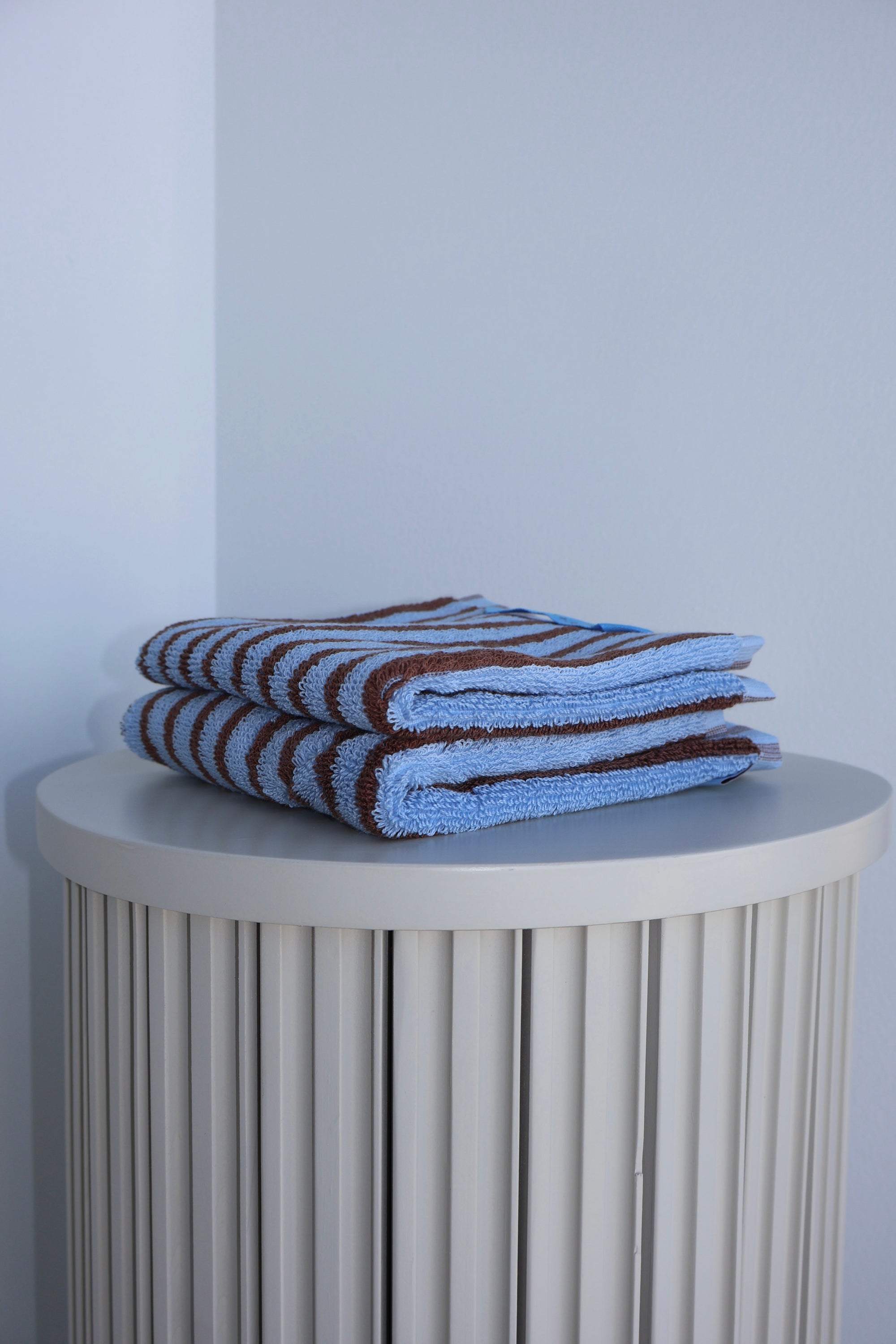 Stack of blue and brown striped towels on a round white table against a light gray wall.