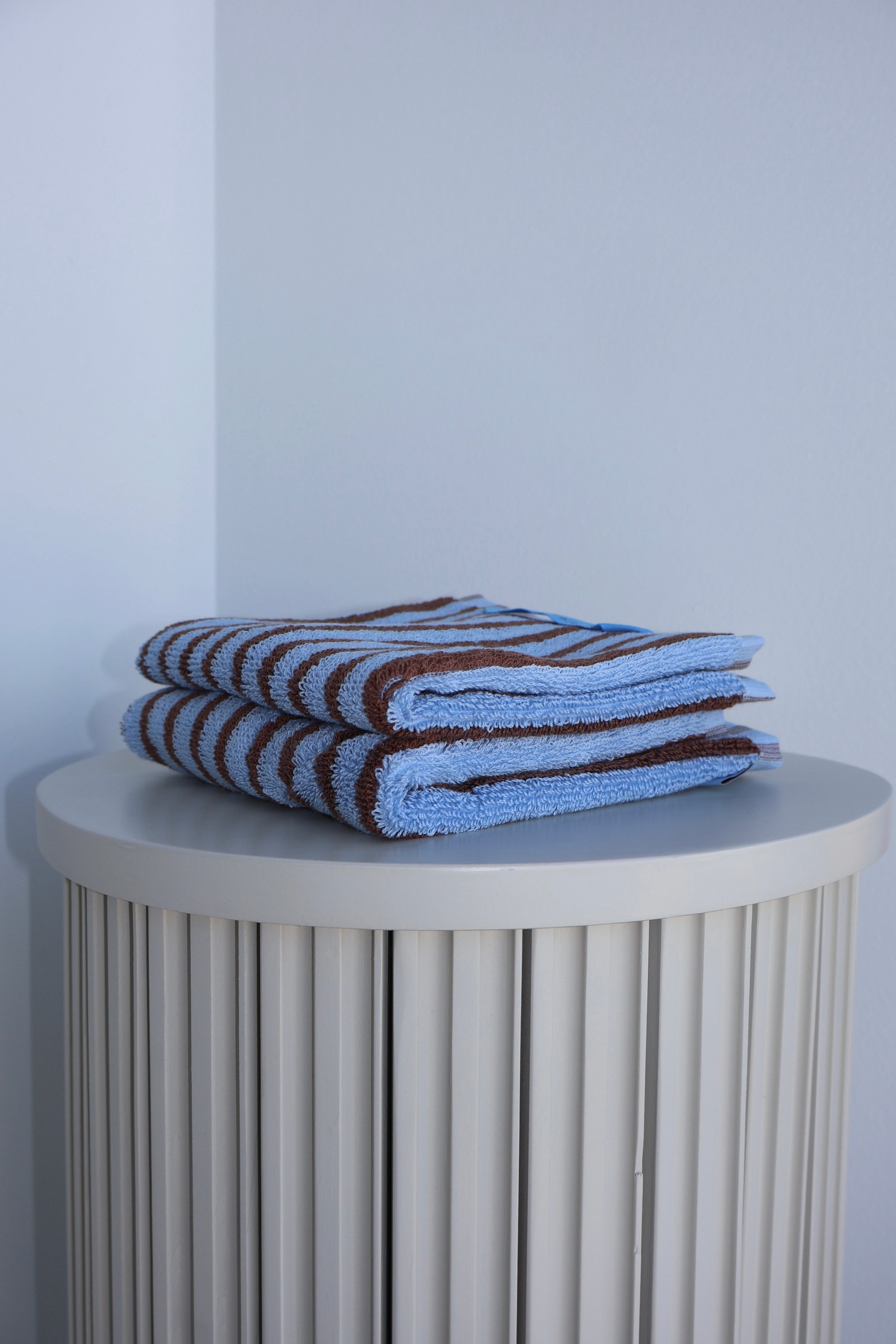 Stack of blue and brown striped towels on a round white table against a light gray wall.